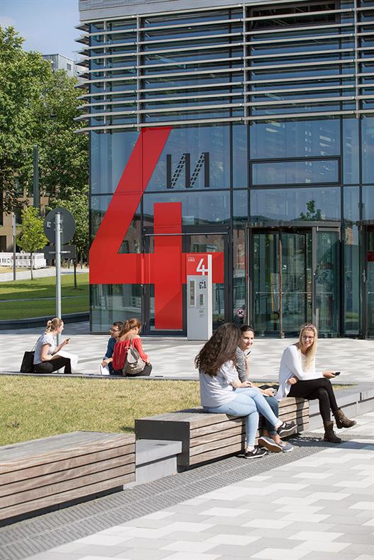 Dieses Bild zeigt das zentrale Hörsaalgebäude der Hochschule Düsseldorf mit einer Fassade aus Stahlblech und Glas. Im Erdgeschoss befinden sich die Hörsäle mit roten Stühlen. Im Obergeschoss gibt es Büroräume für die Beschäftigten der Hochschule.